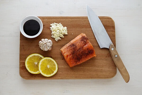 Salmon, lemon slices, garlic, soy sauce, and a chef knife on a wooden cutting board.