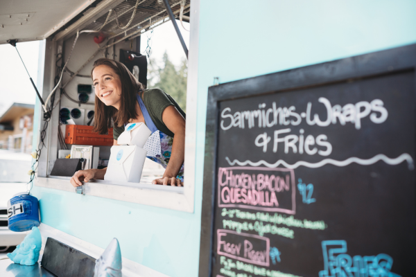 female food truck worker sticks her head out the window smiling and offering a customer their order