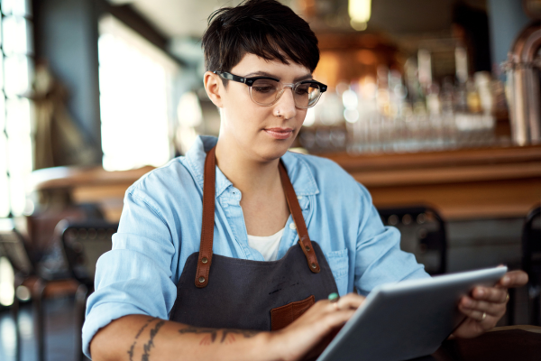 restaurant worker in apron and glasses looks at iPad while standing behind bar