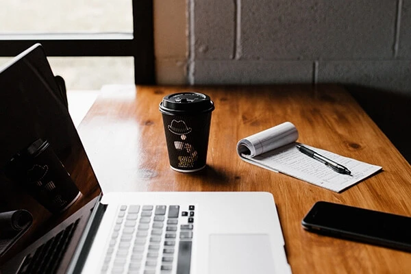 Laptop, coffee cup, paper and pen, and a phone on a wooden table.