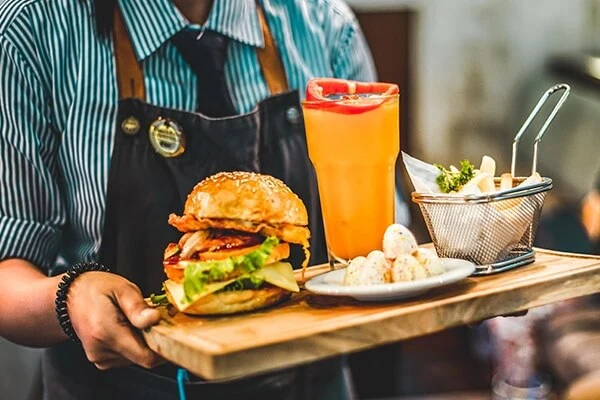 Server carrying a wooden tray with a burger, fries, and a drink out to a diner.
