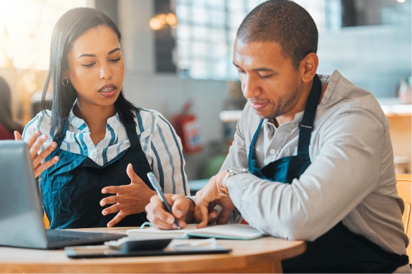 A man and woman discuss at a restaurant table with a laptop, notebook, and pen, engaging in a focused conversation.