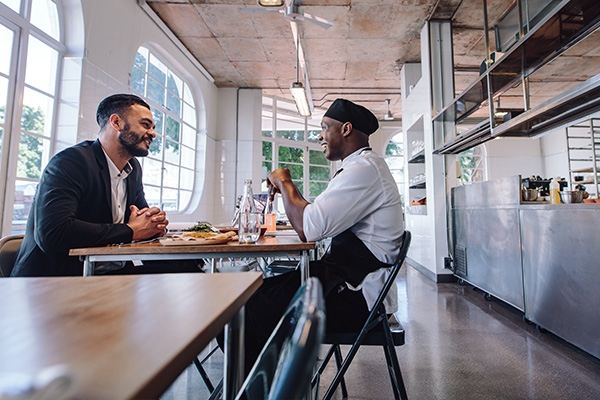 A diner and chef sitting opposite each other at a restaurant table talking.