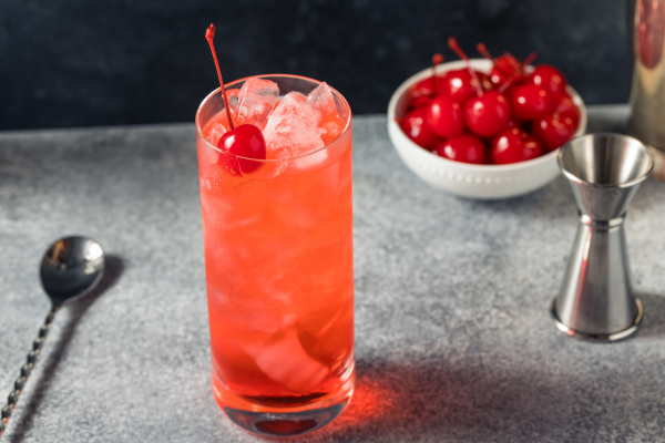 Red cocktail in tall glass over ice with straw and cherry on top, surrounded by bowl of cherries and bar tools